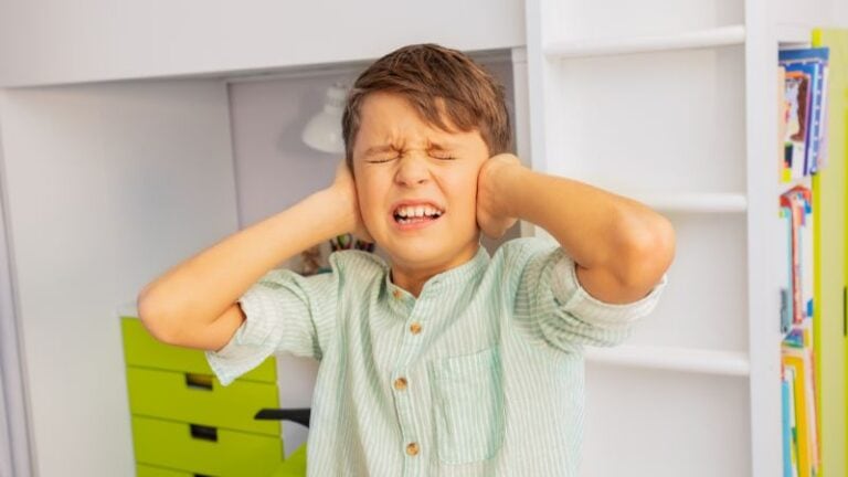 Young boy covering his ears with a distressed expression, standing in a room with white shelves and colorful books, illustrating noise or frustration.