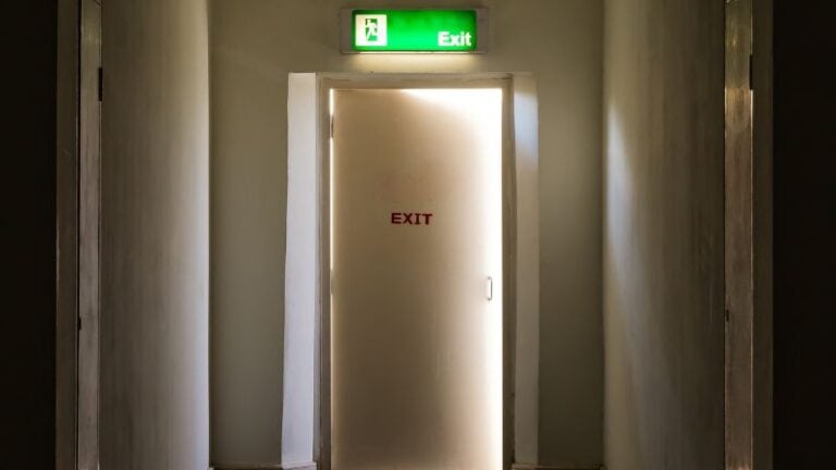 Fire exit door with illuminated green exit sign above, located in a corridor with metallic walls, indicating emergency egress route.